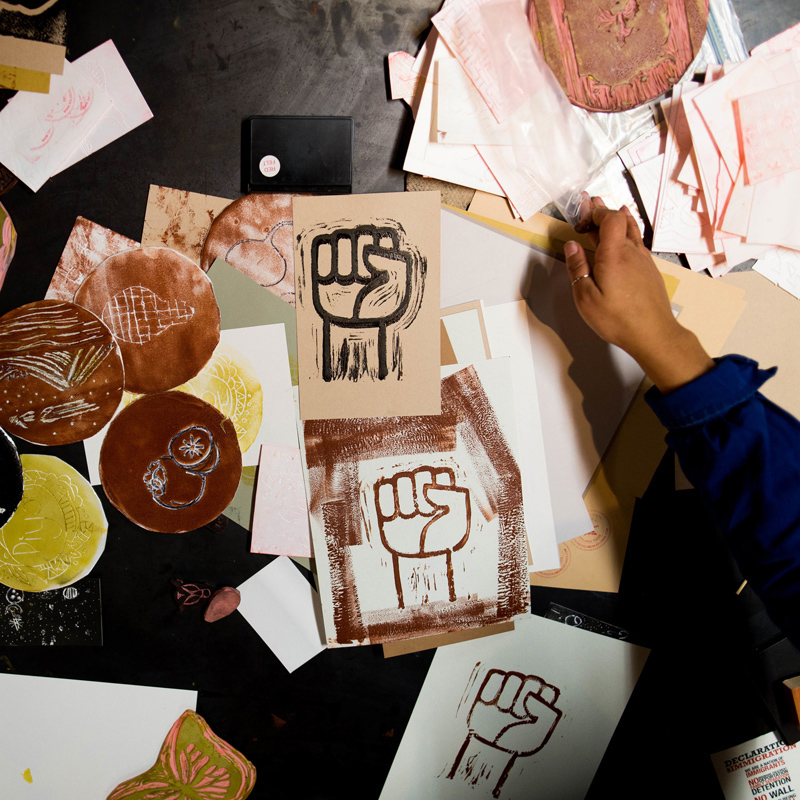 an artist's working space is covered with tortilla-sized print-making plates, and some prints of a hand held into a fist. Image by Faizal Westcott, courtesy of Urbano Project.