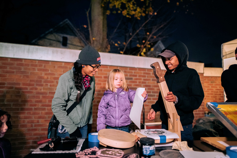 A little girl looks at the print that's come from the torilla press in front of her, while 2 young adults look on, smiling. Image by Faizal Westcott, courtesy of Urbano Project.