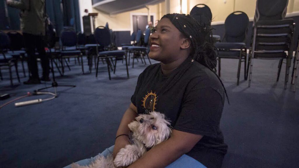 Andrine Pierresaint, sitting on the floor smiling, looking up, a small fluffy dog on her lap looks up at her. Photo: Alexandra Wimley.