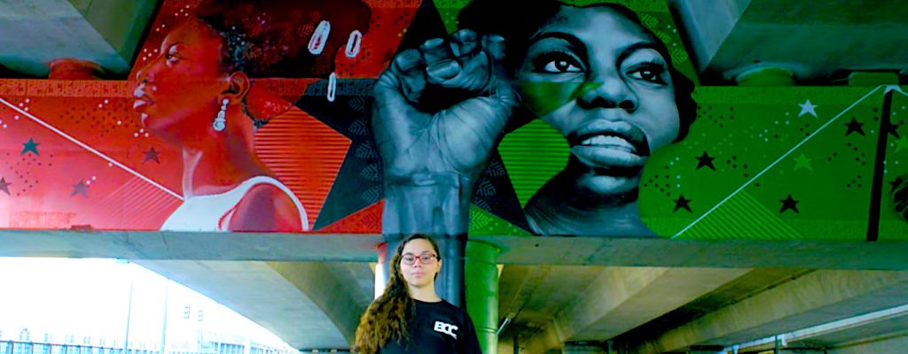 Young woman with long hair and glasses stands in front of a colorful mural with images of black women activists painted on an overpass