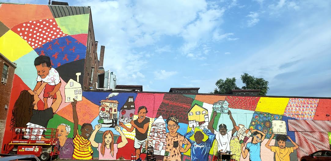 a brightly colored mural depicting young people of different backgrounds holding different kinds of houses (single-family, apartment building). It covers the side of a building. A blue sky above it.